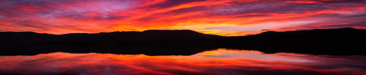 Fototapeta na wymiar Panoramic sunrise or sunset with orange, purple and red clouds reflected in the lake water. Natural mirror at golden hour panorama. Spectacular and beautiful scenery.