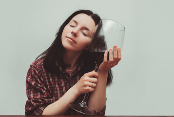 A young girl in a red plaid shirt is tenderly holding a large glass of red wine, closing her eyes. Concept of alcohol abuse, alcoholism, hangover, loneliness and depression. Gray background.