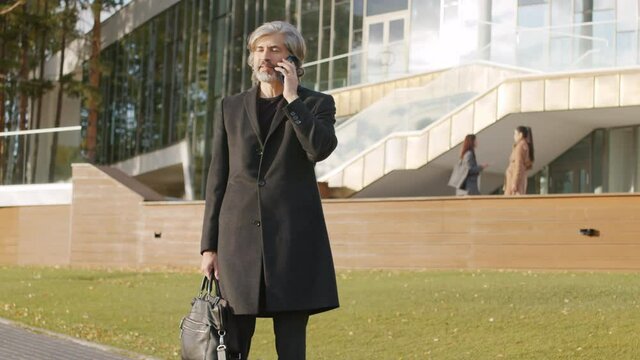 Tilting-up slow-motion footage of mature caucasian man having phone conversation while standing on gyroscooter outdoors in daylight