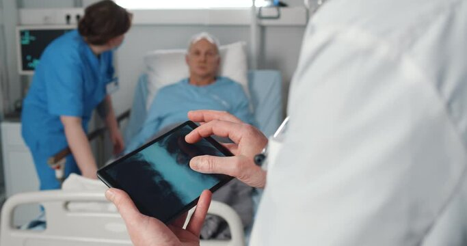 Nurse taking care of aged patient in bed while doctor examining lungs xray on digital tablet