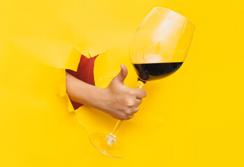 A man's hand emerges through a torn hole in yellow paper with a large glass of red wine. The...
