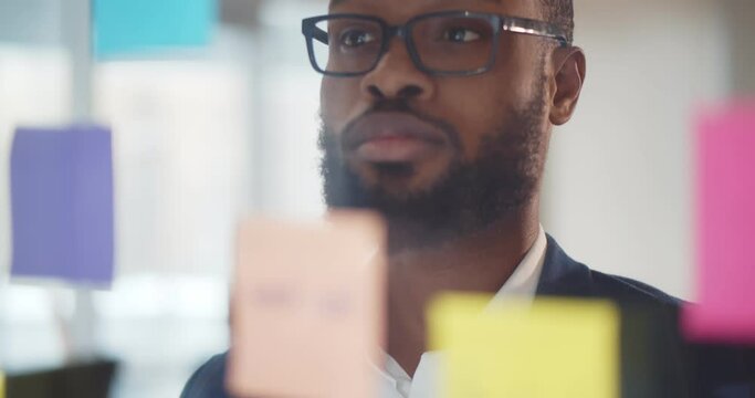 Afro-american businessman writing on sticker with pencil stuck to glass wall.