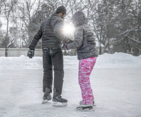 Fototapeta na wymiar Young man teaching girlfriend to skate on outdoor rink with trees and falling sun in background
