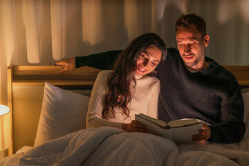 caucasian couple lying on bed reading book in bedroom together