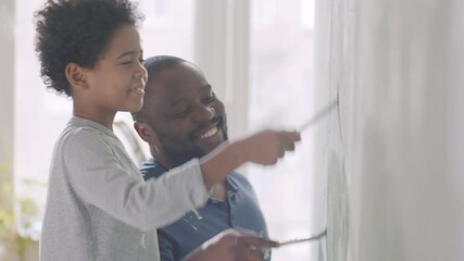 Cheerful African American father and little son smiling and talking while painting wall with brushes during home renovation - Powered by Adobe