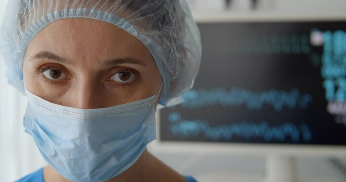 Woman doctor in safety mask and hat with ecg monitor showing patient heart rate on background