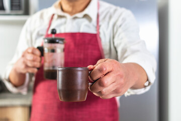 Close-up of a man's hand offering a cup of coffee while holding a French press in the other hand that looks out of focus