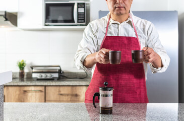 Close-up of an unseen man wearing a red apron offering two cups of coffee at a kitchen counter or...
