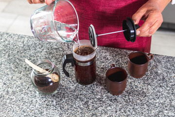 Aerial shot of a barista whose face is not seen preparing two cups of coffee in his kitchen in a French press on a counter.