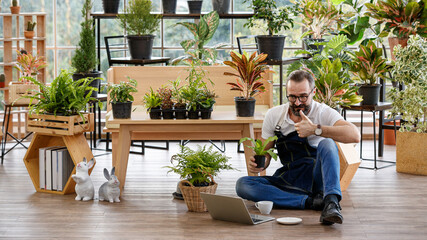 A beautiful bearded man sitting and using laptop notebook computer to sell plant in home indoor...