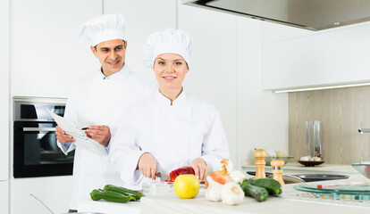 Professional chefs in uniform prepare vegetables with paper recipe on kitchen