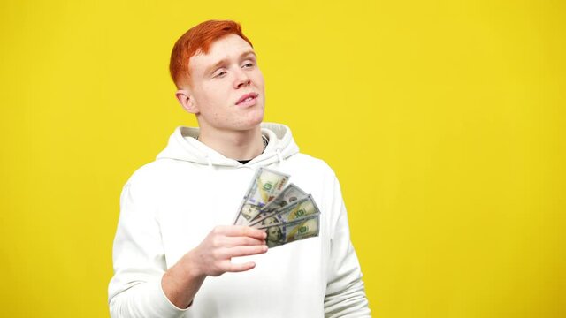 Young redhead man using cash as fan looking at camera with egoistic facial expression. Wealthy Caucasian guy boasting money at yellow background in slow motion.