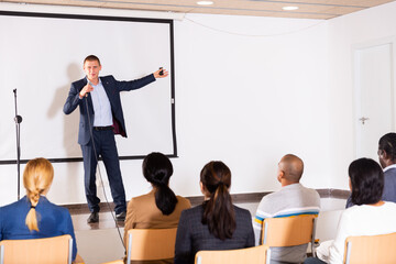 Portrait of expressive inspirational speaker with microphone on stage in conference room during motivational coaching seminar