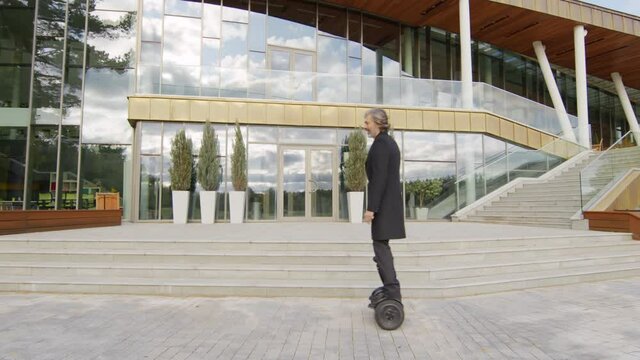 Slow-motion full shot of senior caucasian business-like man riding gyroscooter near modern glass office building in afternoon