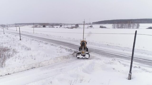 Aerial view of Snowblower Grader Clears Snow Covered Country Road in field next to the forest. The grader drives off the asphalt road into the field. Hard winter weather conditions. Cloudy winter day
