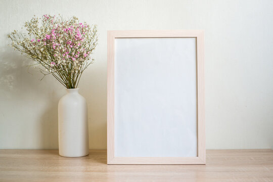 Portrait white picture frame mockup on wooden table. Modern ceramic vase with gypsophila.  White wall background. Scandinavian interior. Vertical. 