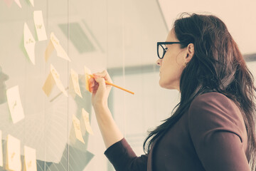 Confident middle-aged businesswoman writing on sticker with pencil and brainstorming. Colorful memo notes stuck on glass wall in conference room. Business, planning and management concept