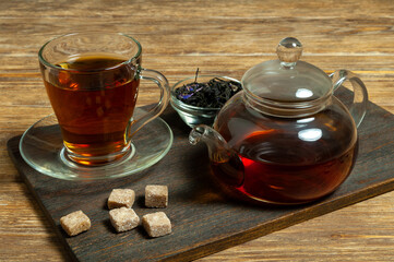 Glass cup of tea with saucer, teapot, tea leaves and brown cane sugar on wooden background. Close-up