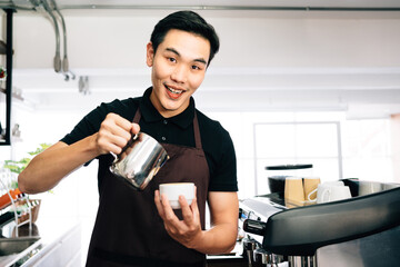 Young Asian male barista looking at the camera, wearing an apron pouring hot milk into hot espresso black coffee for making Latte Art.