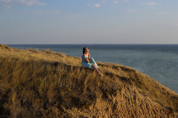 A pretty girl in a long blue dress sits on a high cliff by the sea or ocean on sunny windy summer day. Young beautiful woman with short hair watching sunset on the shores of the calm sea.