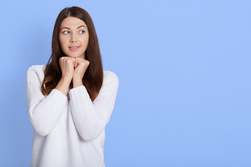 Portrait of cute caucasian woman in white shirt keeps hands under chin, looking aside and sees beautiful, dreams and thinks about something pleasant, posing isolated over blue background.