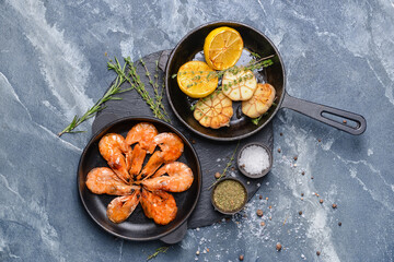 Frying pans with tasty shrimps, garlic and lemons on color background