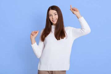 Fototapeta na wymiar Portrait of optimistic dark haired young woman poses with hands up, dances carefree, wears white jumper, looks at camera, isolated over blue wall, winsome lady celebrates happily.