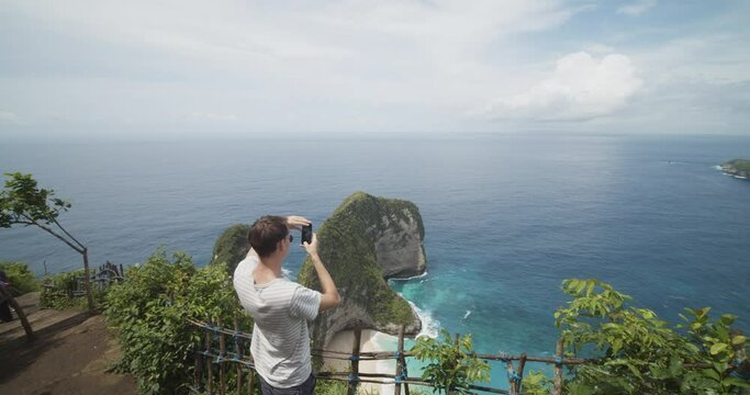 Back ascending view of a male tourist taking pictures of the T-Rex Bay peninsula at Kelingking beach in Bali, Indonesia with his phone Man at the tropical beach observation point