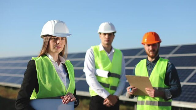 The female engineer is standing and looking at the camera. Two engineers are also looking at the camera. They are standing next to solar panels. 4K.