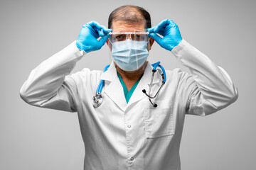 Middle-aged male doctor putting on face shield, grey background