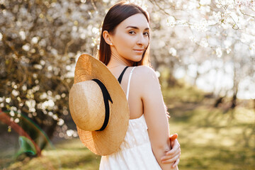 Girl with a straw hat in the spring in the park. Brunette with long hair holds a hat on a background of summer nature. Youth and beauty.