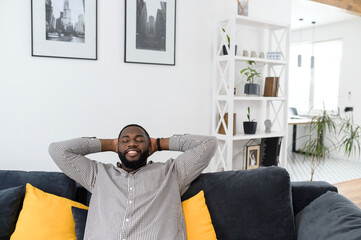 Satisfied young african man resting on comfortable couch in white cozy modern living room, holding...