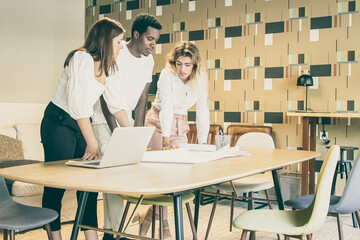 Obraz premium Content multiethnic designers standing and looking at draft. Concentrated professional colleagues meeting in coworking space, standing and discussing project. Communication and design concept