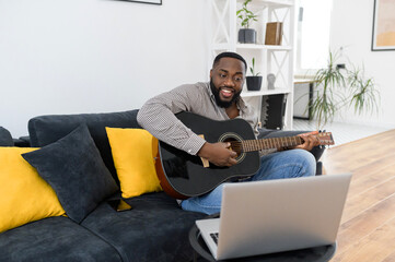 Smiling young African American male musician playing guitar during an online concert at home while...