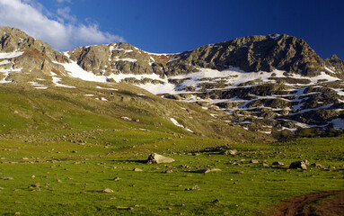 A panoramic shot of a mountain filled with snow behind a green path which has stones on it under...