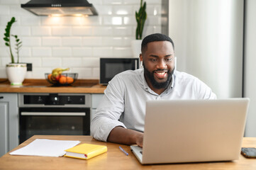 Smiling African American guy sitting at the table at home in the modern kitchen, using the laptop for distance video communication, studying, working, meeting online, chatting, browsing the internet