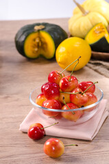 Fresh red cherries in glass bowl with pastel pink tablecloth on the wooden table and vegetable in background