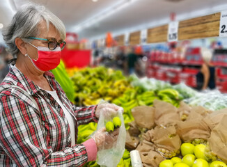Grey-haired senior woman wearing a red surgical mask due to coronavirus looking and choosing fruits in a supermarket