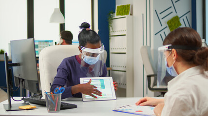 Multiethnic businesswomen discussing work and using digital tablet during meeting wearing protection mask respecting social distance. Group of diverse businesspeople working in background.