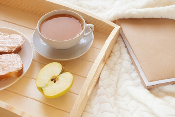 Wooden tray with a cup of coffee and an apple on a light blanket. There is a book nearby. View from...