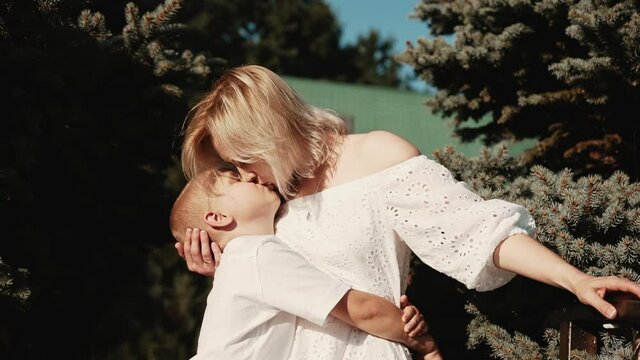 Joyful mother and son spend the weekend together and hug near coniferous trees and kiss expressing their love