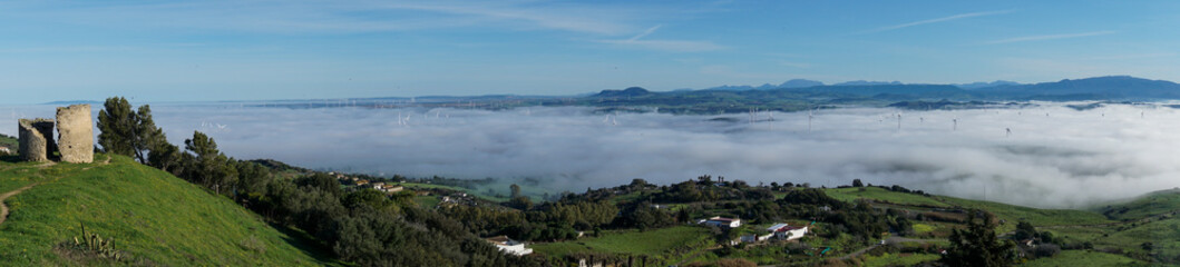 panorama view of rolling hills landscape in Andalusia with many wind turbines above the fog in the valleys and blue sky above