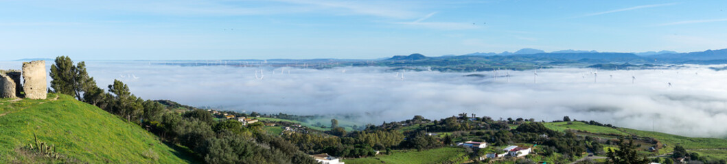 panorama view of rolling hills landscape in Andalusia with many wind turbines above the fog in the valleys and blue sky above