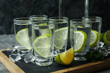 Board with bottle and shots of vodka with lime on gray table