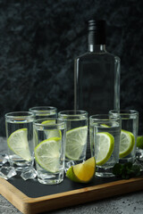 Board with bottle and shots of vodka with lime on gray table