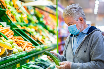Senior adult man with surgical mask due to coronavirus while shopping at the supermarket. White-haired retiree while he chooses zucchini in the vegetable department. Consumerism concept