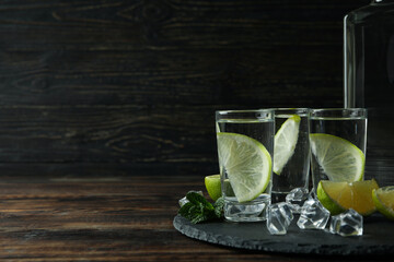 Tray with bottle and shots of vodka, lime and ice on wooden background