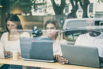 Three designers working on laptops behind window with reflection. Serious multiethnic workers sitting at table and using portable computers. Communication and digital technology concept