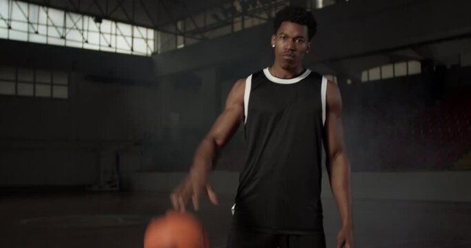 Video portrait of a basketball player bouncing a ball. African american man in uniform posing inside sports hall