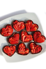 Fototapeta premium Valentine’s day dessert background. Mini heart shaped chocolate tarts with fresh strawberries on a plate isolated on white